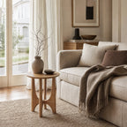 Elin Oak Side Table with a circular top and sculptural base in natural oak, beside a beige sofa in a sunlit living room.