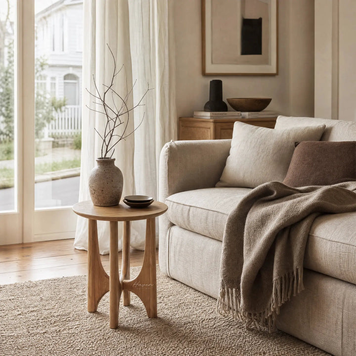 Elin Oak Side Table with a circular top and sculptural base in natural oak, beside a beige sofa in a sunlit living room.