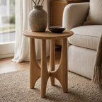 Elin Oak Side Table in natural oak with a sculptural base and circular top beside a sofa, speckled vase.