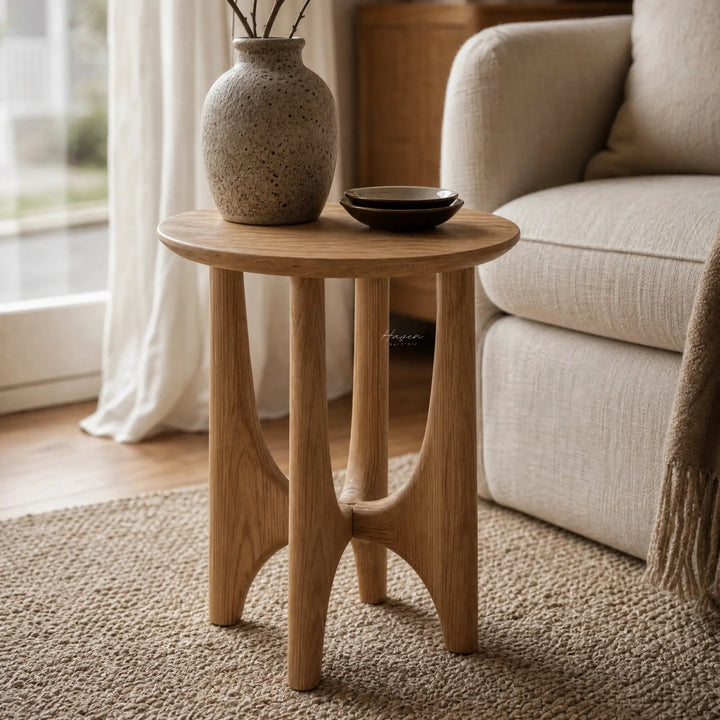 Elin Oak Side Table in natural oak with a sculptural base and circular top beside a sofa, speckled vase.