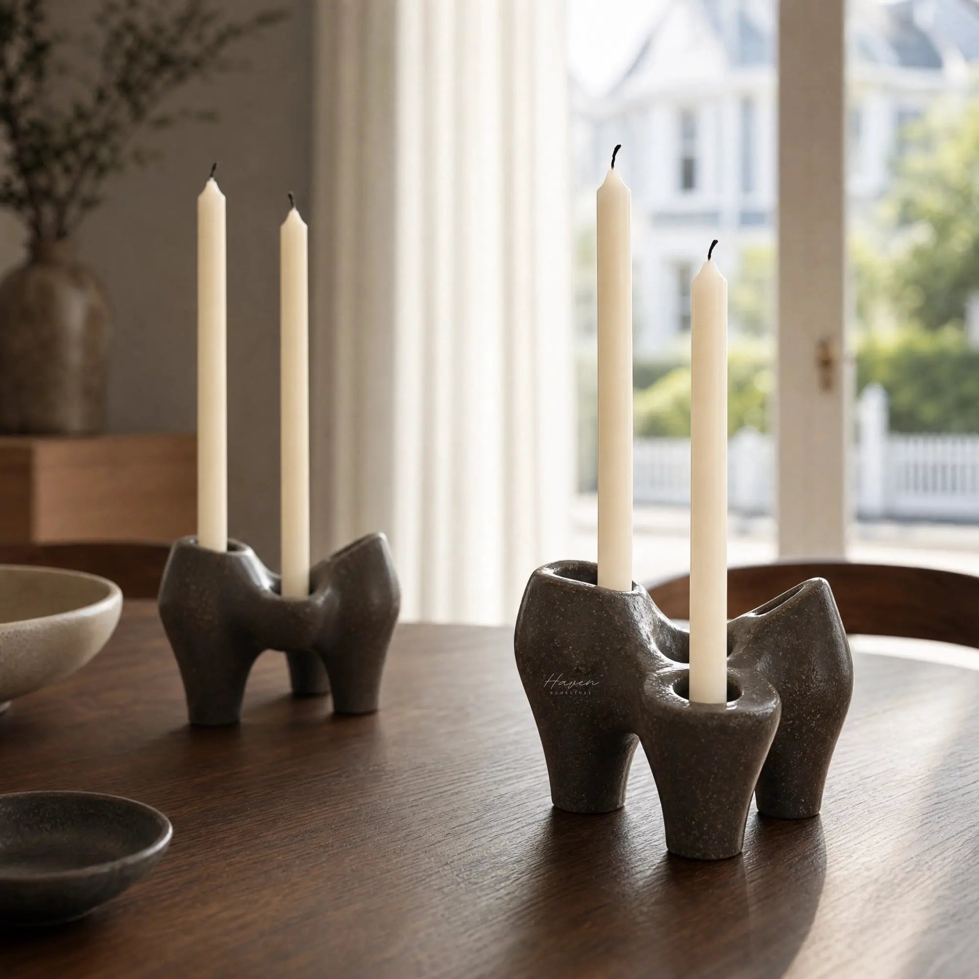 Broste Laia candleholder set/2 in antique brown glaze with tall taper candles on a wooden table by a window.