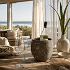 MUUBS Valley Wine Cooler in riverstone on a rustic wooden table with wine and glasses, seaside setting.