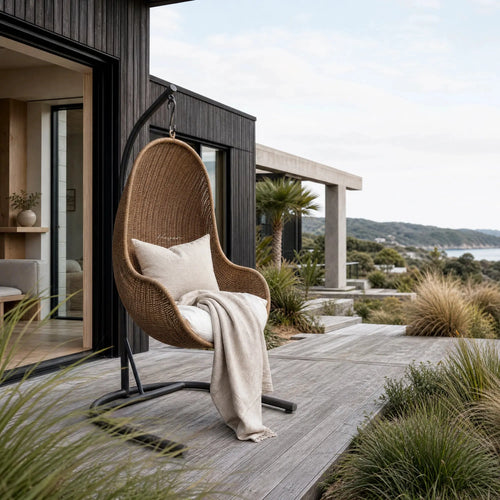 Cabana Pod Chair hanging wicker seat with cushions on a wooden deck beside a modern house.