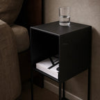 STUDIO Bedside Cabinet Small in black metal with an open shelf and a glass of water on top.