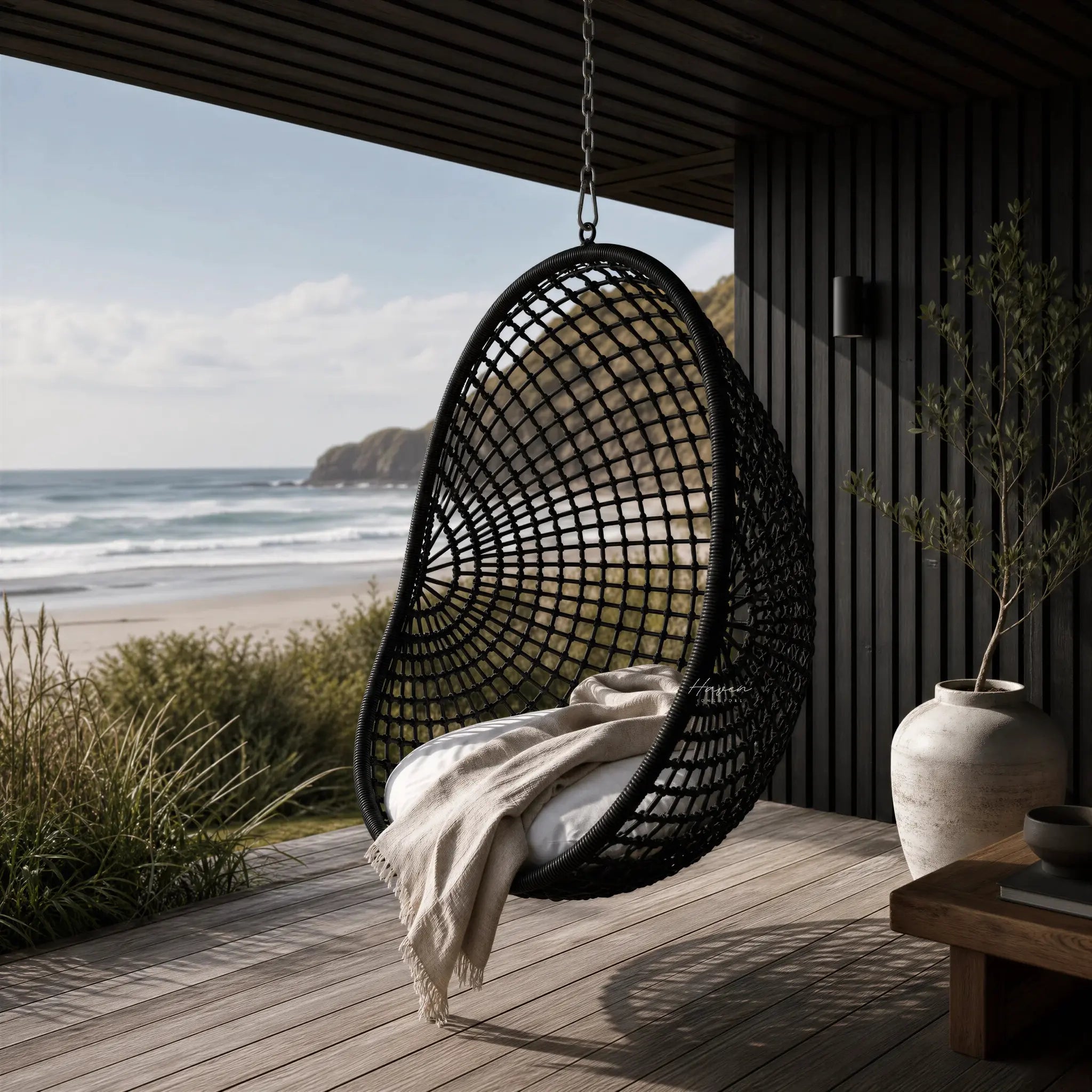 Check Pod Chair hanging on a deck with coastal view, woven black mesh over galvanised metal frame, and beige blanket.