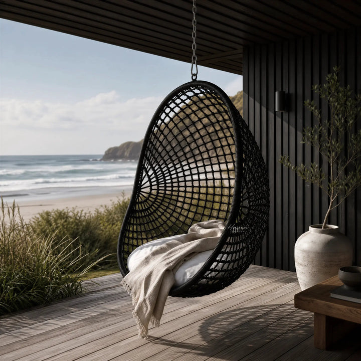 Check Pod Chair hanging on a deck with coastal view, woven black mesh over galvanised metal frame, and beige blanket.