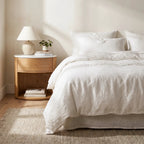 Dana Bedside Table with warm oak, marble top, and rounded silhouette beside a bed.