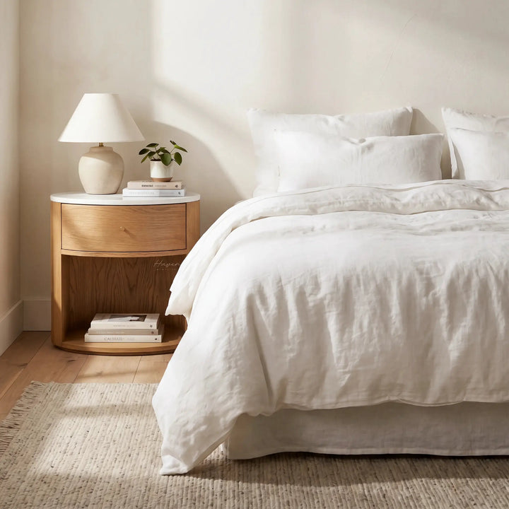Dana Bedside Table with warm oak, marble top, and rounded silhouette beside a bed.