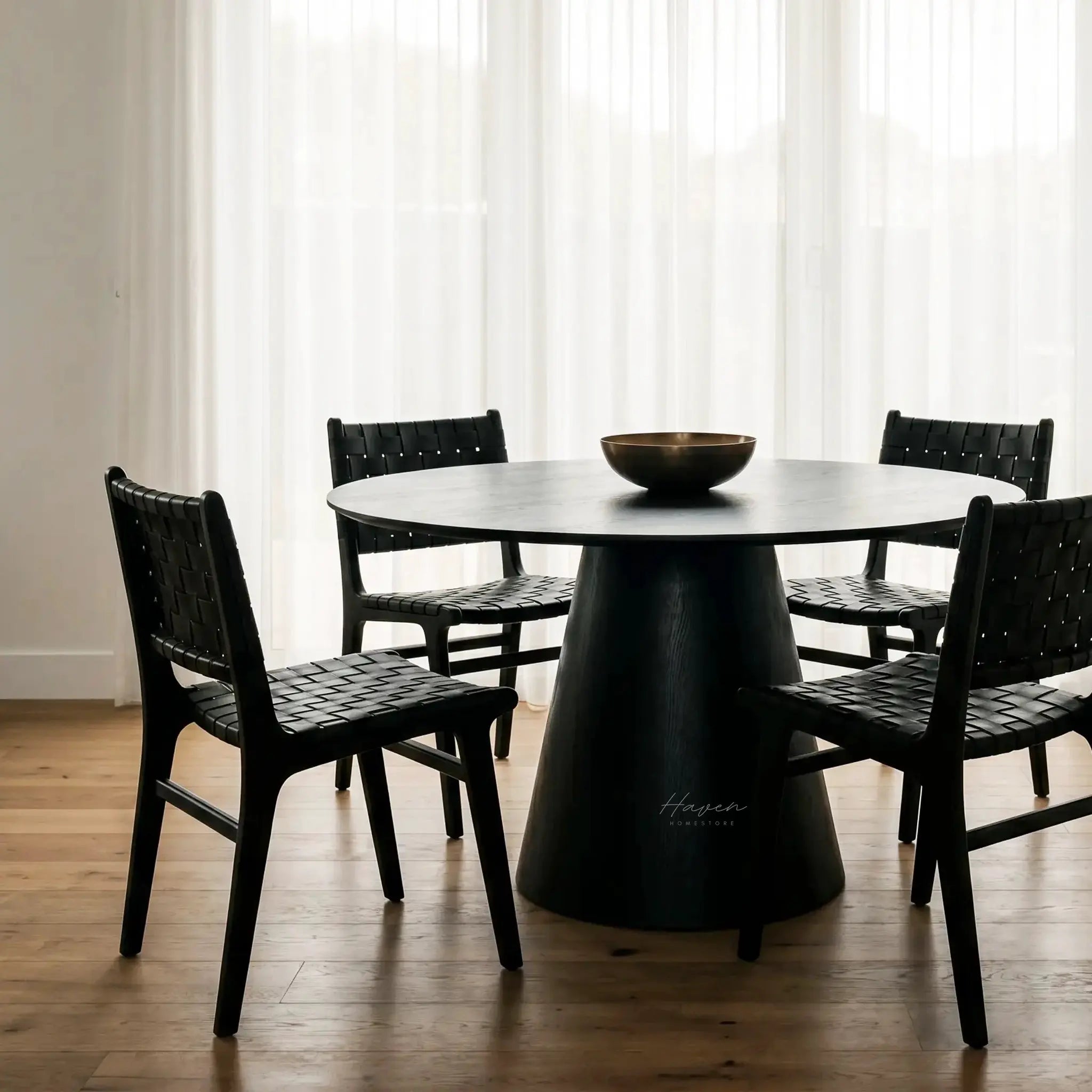 Charlie Dining Table with a bold pedestal base and rounded top in black oak veneer, surrounded by black woven chairs.
