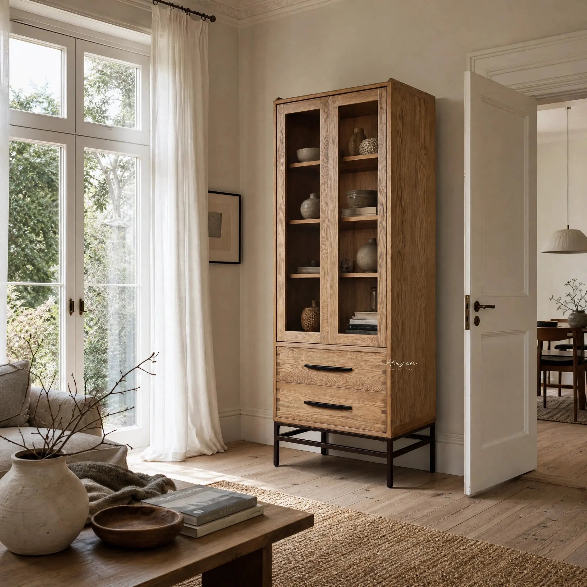 Halden Display Cupboard in solid oak with glass-front doors, showcasing ceramics in a bright living room.