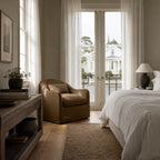 Softly curved Nella slipcover chair in warm Mocha linen positioned in a sunlit bedroom beside sheer curtains.