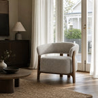 Seki Chair in Pumice fabric features a curved upholstered back and exposed solid ash frame positioned near a window with sheer curtains.