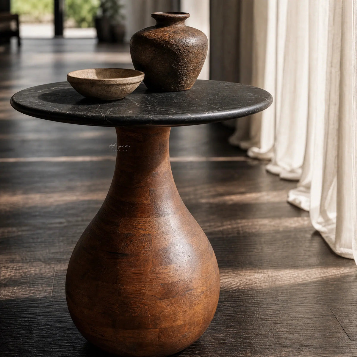 Sculptural side table with black marble top and tapering mango wood pedestal base, displaying rustic ceramic vessels in natural light.