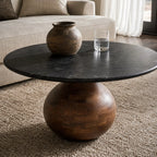 Round black marble coffee table with a sculptural rounded mango wood pedestal base, styled with a ceramic vase and glass of water on a beige rug.
