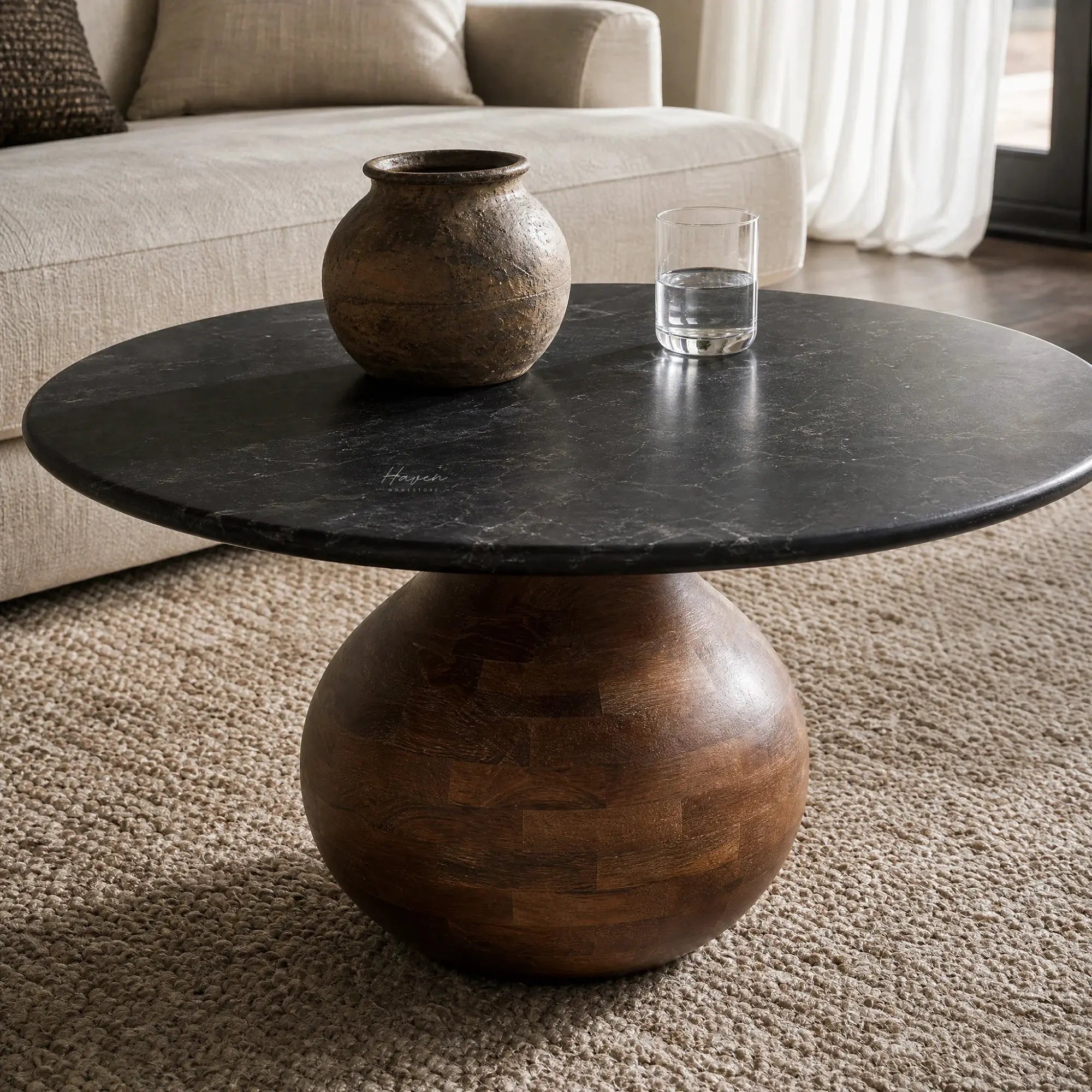 Round black marble coffee table with a sculptural rounded mango wood pedestal base, styled with a ceramic vase and glass of water on a beige rug.
