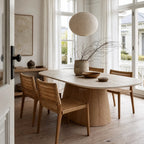 Oval faux marble dining table with a tapered wooden pedestal base and woven chairs in a sunlit room.