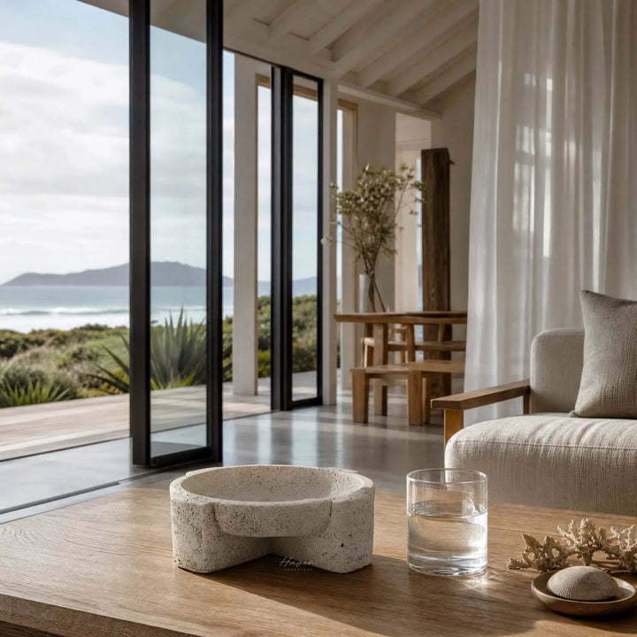 Textured concrete bowl with irregular form on wooden table in coastal living room featuring ocean view.