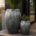 Set of two rounded fibre cement planters with textured black-grey volcanic finish, holding greenery on a wooden deck.