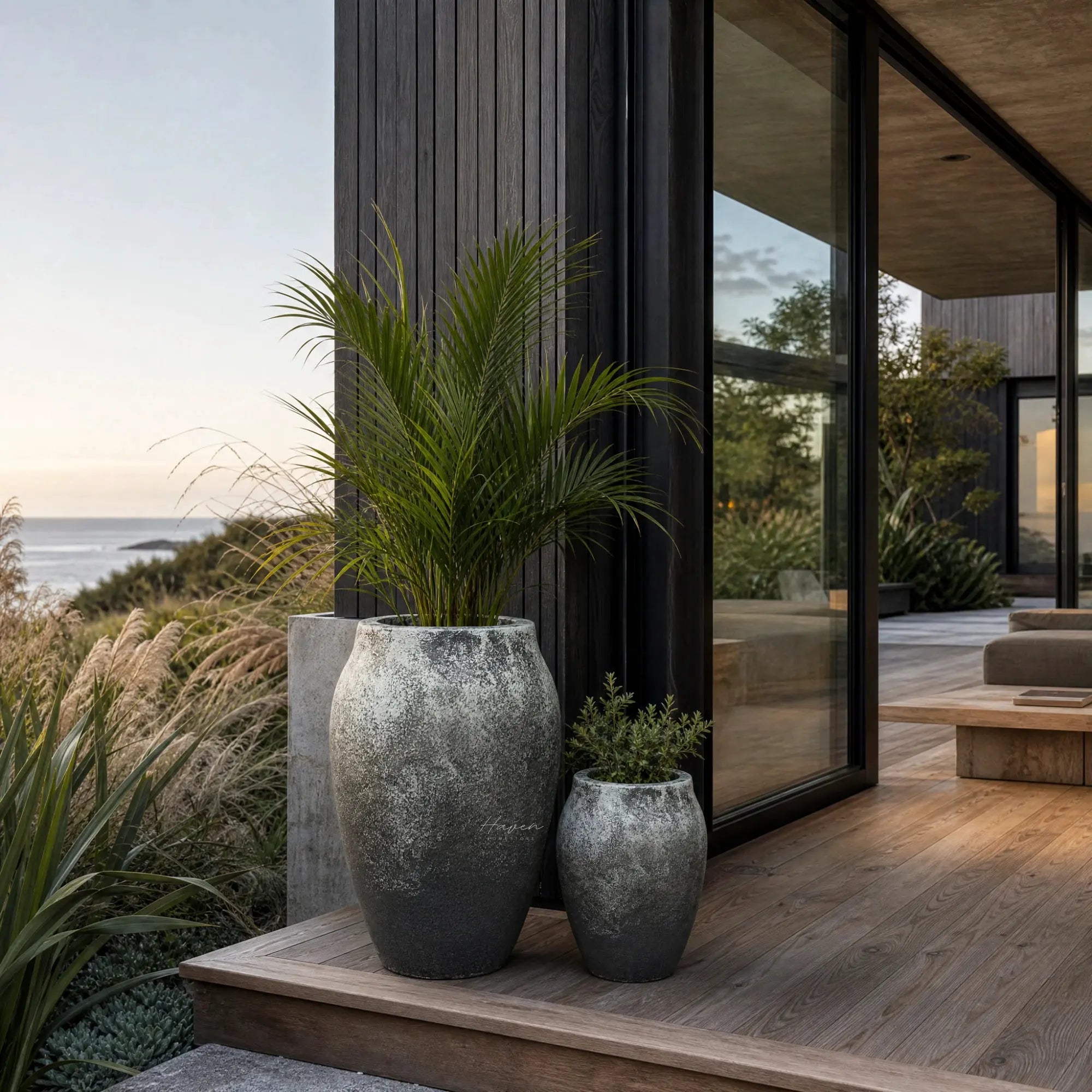 Textured black-grey fibre cement lava planters set displaying rounded forms on a wooden deck beside modern architecture and ocean view.