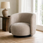 Oatmeal swivel chair with rounded profile, textured polyester upholstery, and dark base beside wooden side table and lamp.