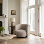 Oatmeal swivel chair with rounded profile and textured fabric, grounded by a dark base in a sunlit living room.