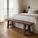 Rustic reclaimed elm bench with substantial legs at the foot of a bed, featuring visible knots and grain.