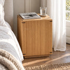 Warm oak side table with faceted design and dark corner accents sits beside a bed holding a magazine and water glass.