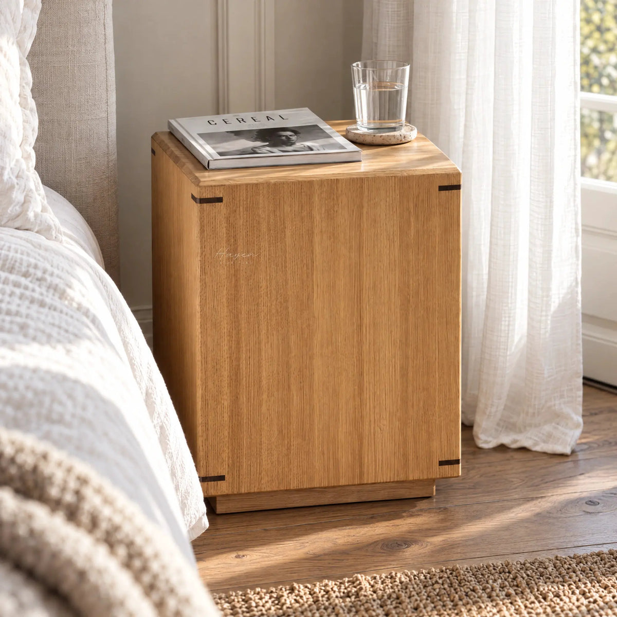 Warm oak side table with faceted design and dark corner accents sits beside a bed holding a magazine and water glass.