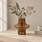 Tabac Vase holding dried foliage. Sculptural amber glass vase features a low, rounded body, narrow neck, and cylindrical base on a stone surface.