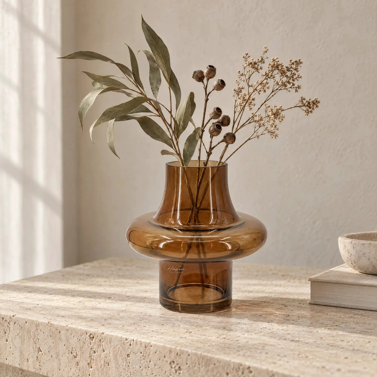 Tabac Vase holding dried foliage. Sculptural amber glass vase features a low, rounded body, narrow neck, and cylindrical base on a stone surface.