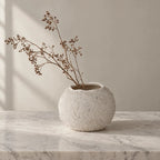 Textured round off-white vase with irregular rim holding dried branches on a marble surface.