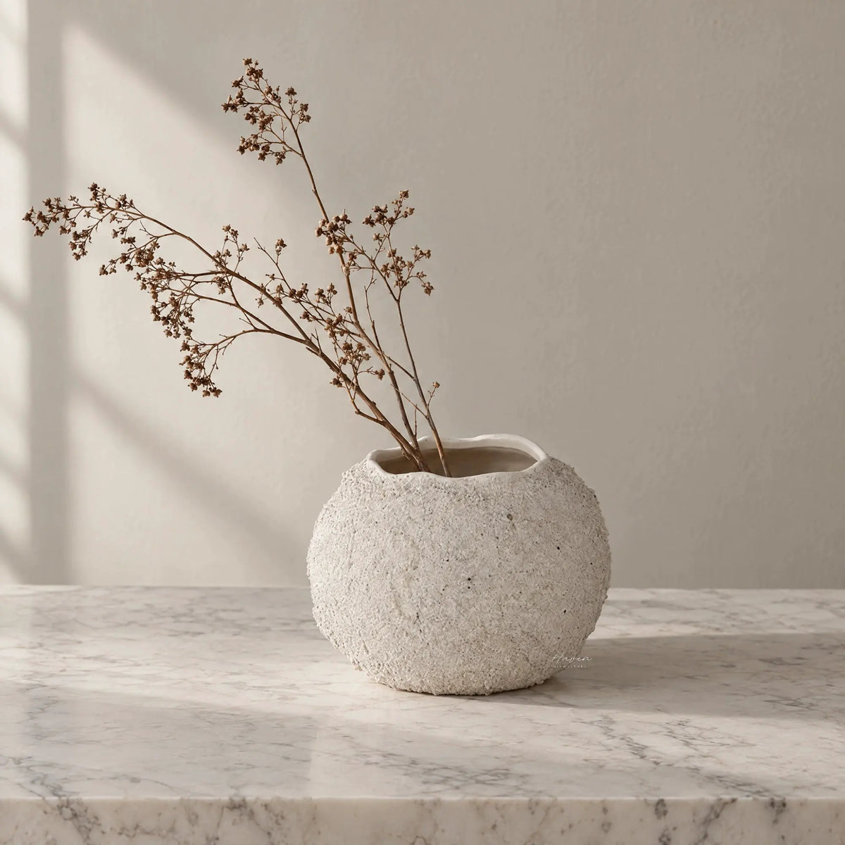 Textured round off-white vase with irregular rim holding dried branches on a marble surface.