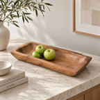 Solid teak tray with organic rectangular shape and visible grain holds green apples on marble surface next to ceramic vase and books.