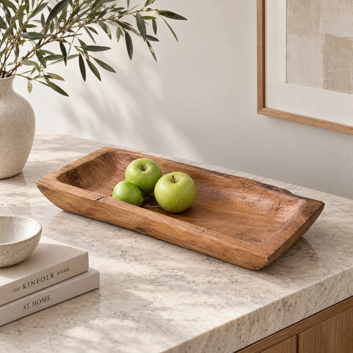 Solid teak tray with organic rectangular shape and visible grain holds green apples on marble surface next to ceramic vase and books.