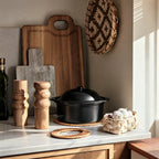 Natural oiled oak salt and pepper grinders with a sculpted three-ring profile sit on a marble counter alongside a black pot, cutting boards, and garlic bulbs.
