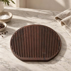 Round charred ash cheese board with linear grooves resting on marble, accompanied by salt and pepper bowls.