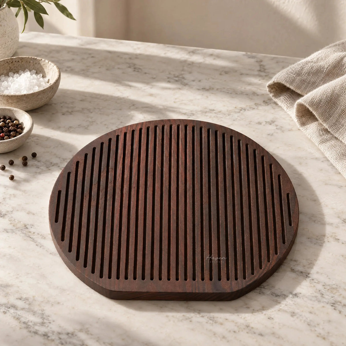 Round charred ash cheese board with linear grooves resting on marble, accompanied by salt and pepper bowls.