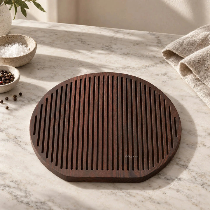 Round charred ash cheese board with linear grooves resting on marble, accompanied by salt and pepper bowls.