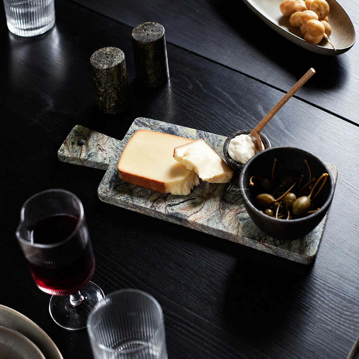 Green marble serving board with integrated handle displays cheese, capers, and dip on a dark wooden table setting.