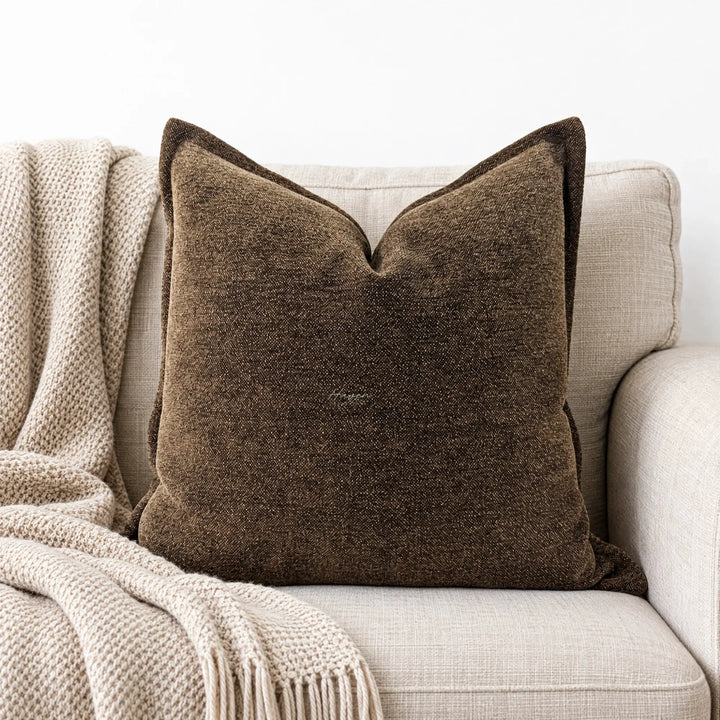 Textured cocoa cushion cover with subtle tonal variation resting on a beige sofa beside a chunky knit throw.