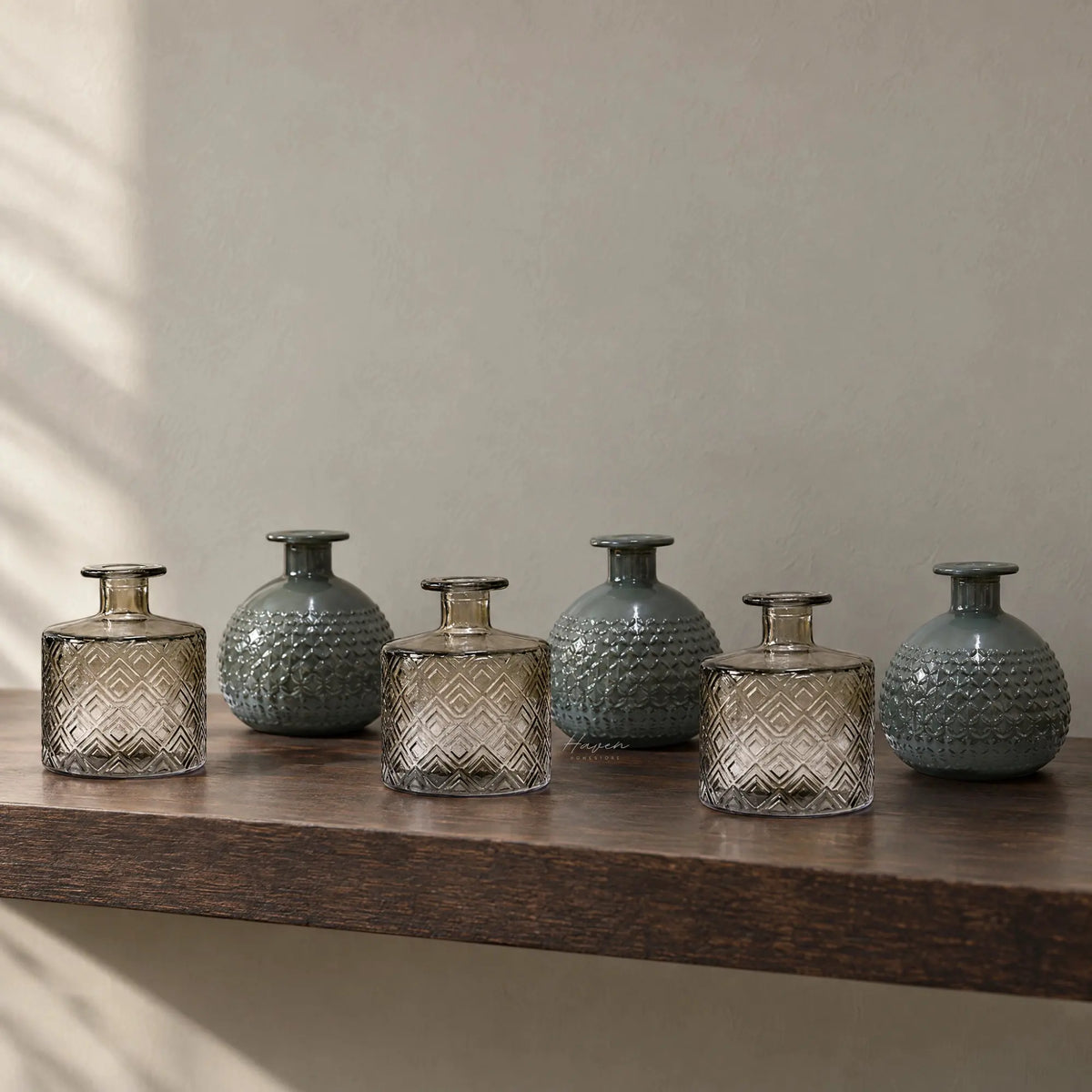 Set of six Spanish glass vessels in smoky grey and teal. Alternating cylindrical and rounded shapes feature embossed geometric textures displayed on a wooden shelf.
