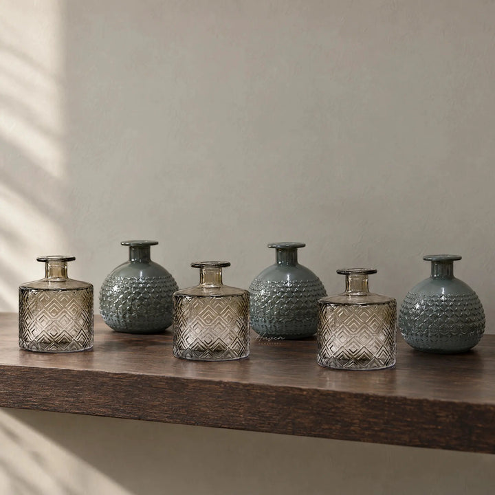 Set of six Spanish glass vessels in smoky grey and teal. Alternating cylindrical and rounded shapes feature embossed geometric textures displayed on a wooden shelf.