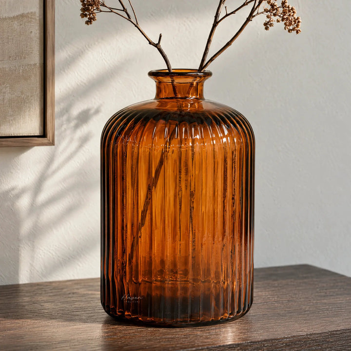 Amber glass vase with vertical fluting and rounded shoulders holding dried branches on a wooden surface.