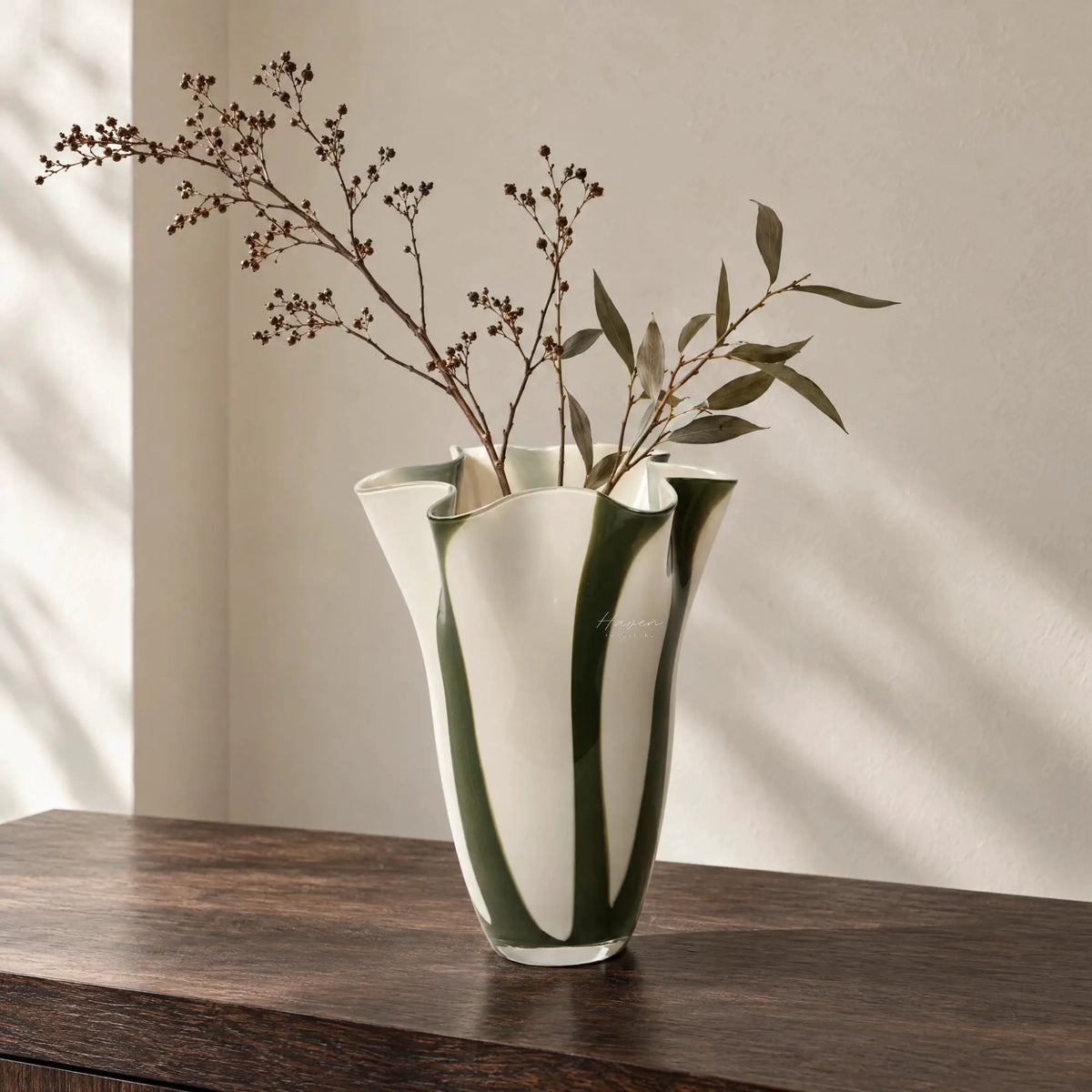 Sculptural glass vase with fluid silhouette featuring vertical strokes of hunter green and soft cream white on a wooden surface.