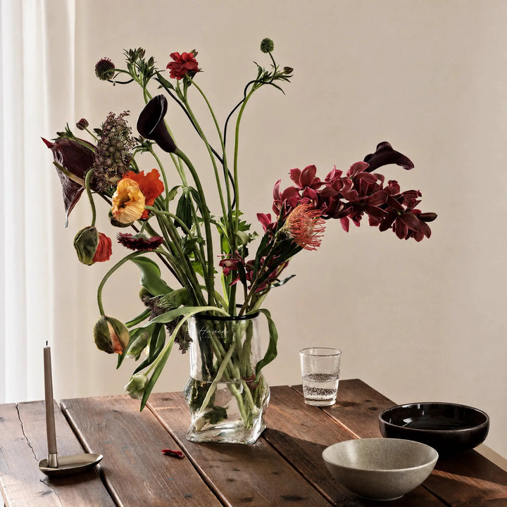 Clear mouthblown glass vase with a black rim and sculptural folded base holding a wildflower arrangement on a wooden table.