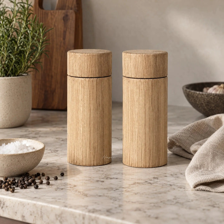 Two cylindrical natural oak salt and pepper grinders standing on a marble countertop beside a bowl of coarse salt and loose black peppercorns.