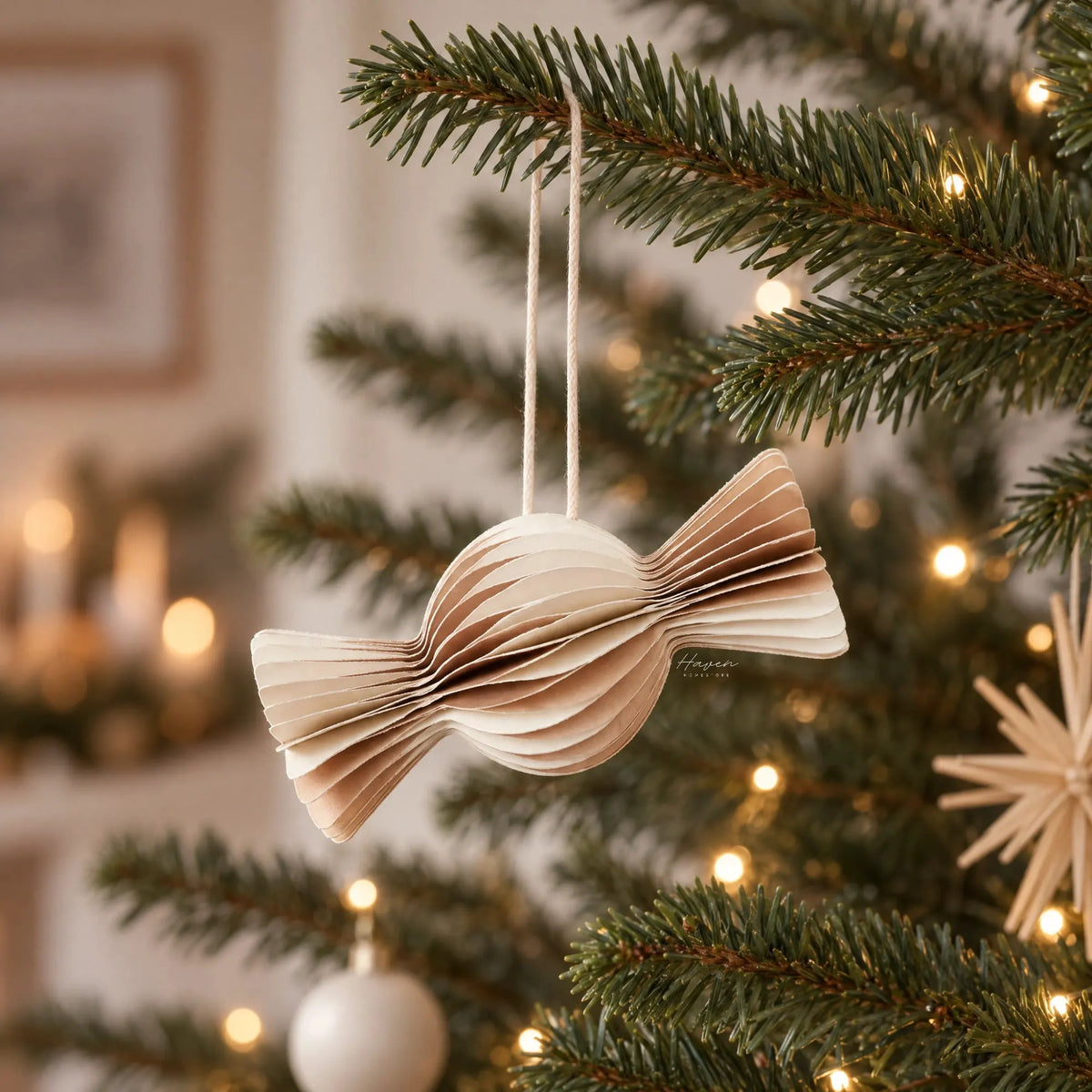 Paper candy ornament with layered off-white and brown folds hanging on a Christmas tree branch with warm lights.