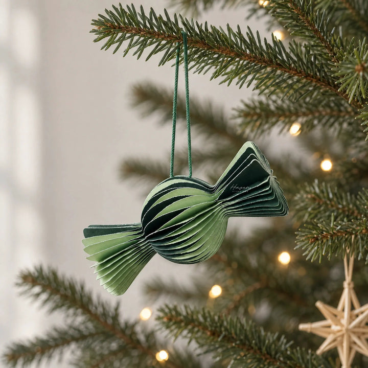 Layered paper candy ornament in alternating light and forest green hangs from a pine branch with warm lights.