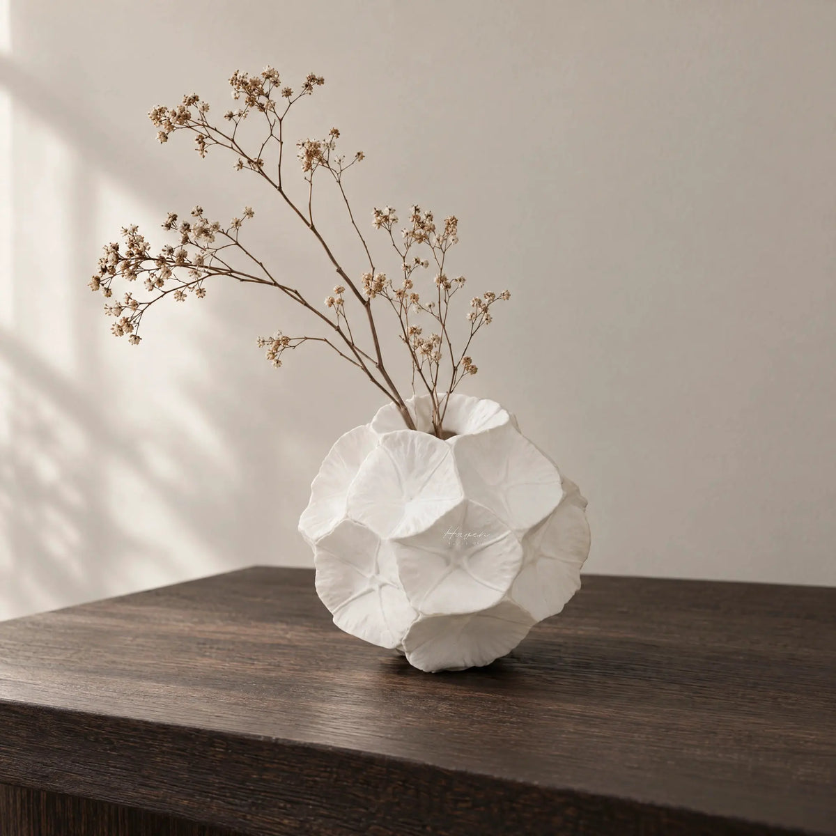White bone china star-studded seed pod vase with petal-like impressions holding dried branches on a dark wood table.