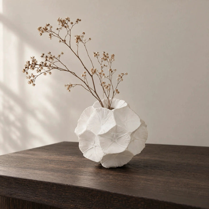 White bone china star-studded seed pod vase with petal-like impressions holding dried branches on a dark wood table.